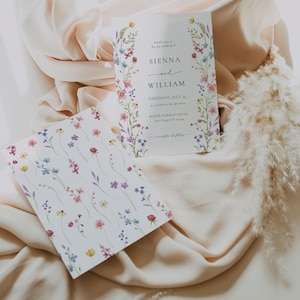 Puede incluir: Invitaci&oacute;n de boda con borde floral y tarjeta a juego. La invitaci&oacute;n dice "Sienna y William" con detalles del evento. La segunda tarjeta tiene un patr&oacute;n floral. Ambas tarjetas est&aacute;n sobre una tela color crema.