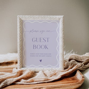 May include: A white guest book sign with a purple border and text. The sign says 'Please sign our Guest Book' and 'Kindly leave your name and wishes for the happy couple.' There is a small purple heart at the bottom of the sign.
