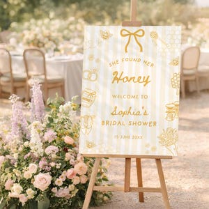 May include: A wooden easel displays a sign with the text "She found her Honey" and "Sophia's Bridal Shower." The sign has a cream background with vertical stripes and gold illustrations of bees, honeycombs, and wedding rings. A floral arrangement is in the foreground.