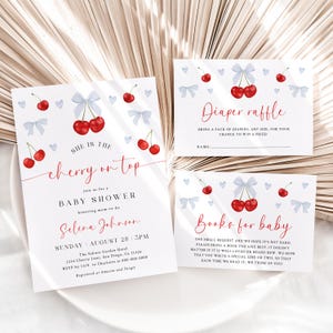 May include: A baby shower invitation with a cherry theme. The invitation is white with red cherries and blue bows. The text reads "She is the cherry on top" and includes details about the shower, including the date, time, and location. There are also two cards for guests to bring diapers or books for the baby.