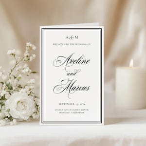May include: A folded wedding invitation with a black border and elegant script. The text reads "Aveline and Marcus" with the date and location. White flowers and a lit candle are in the background.