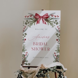 May include: A welcome sign for a bridal shower, featuring a red bow and floral accents. The sign reads "Welcome to Amelia's Bridal Shower" with the date "24 November 20XX". The sign is displayed on a wooden easel.