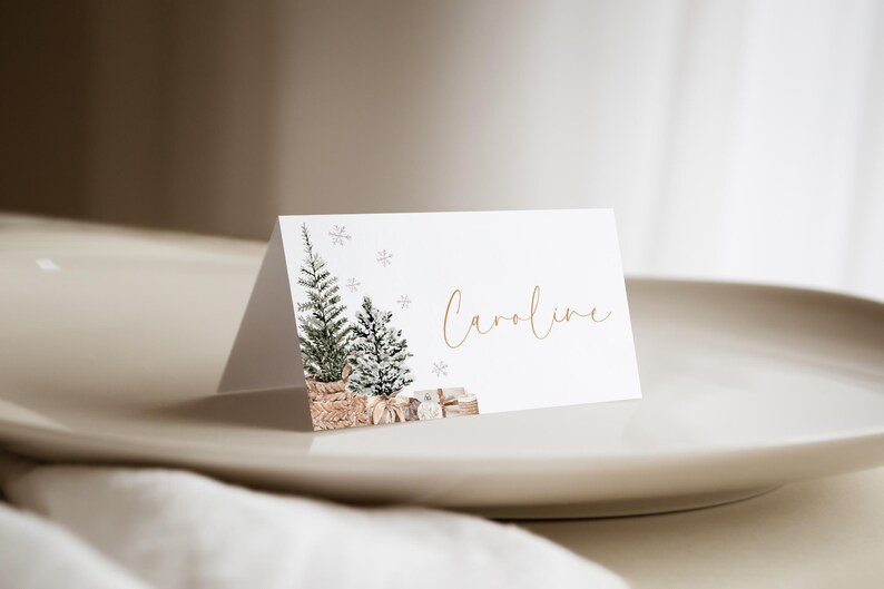 Puede incluir: Una tarjeta de sitio blanca con una ilustraci&oacute;n de acuarela de dos &aacute;rboles de Navidad y copos de nieve. El nombre "Caroline" est&aacute; escrito en escritura dorada. La tarjeta est&aacute; sobre un plato blanco.
