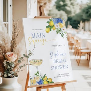 May include: A white sign with blue and yellow floral illustrations and the text "She found her Main Squeeze" and "Welcome to Sophia's Bridal Shower" with the date and location of the event.