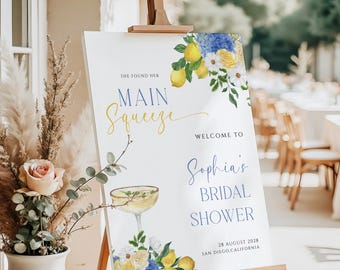 May include: A white sign with blue and yellow floral illustrations and the text "She found her Main Squeeze" and "Welcome to Sophia's Bridal Shower" with the date and location of the event.