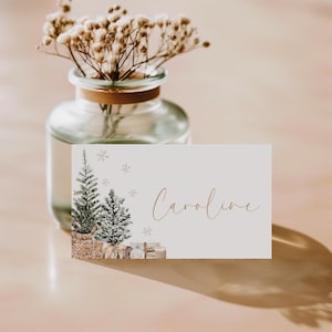 Puede incluir: Una tarjeta de mesa con tem&aacute;tica navide&ntilde;a con el nombre "Caroline" en escritura dorada. La tarjeta presenta ilustraciones de acuarela de dos &aacute;rboles de Navidad, regalos y copos de nieve. Un jarr&oacute;n de cristal con flores secas est&aacute; en el fondo.