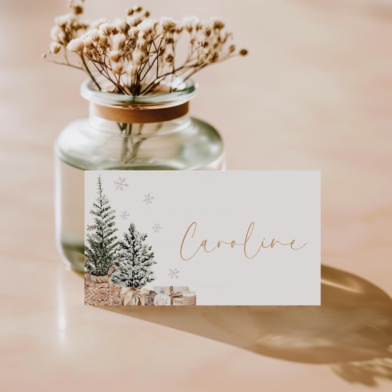 Puede incluir: Una tarjeta de mesa con tem&aacute;tica navide&ntilde;a con el nombre "Caroline" en escritura dorada. La tarjeta presenta ilustraciones de acuarela de dos &aacute;rboles de Navidad, regalos y copos de nieve. Un jarr&oacute;n de cristal con flores secas est&aacute; en el fondo.