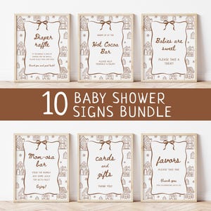 Könnte beinhalten: Ein Set von zehn Babyparty-Schildern mit Lebkuchenhaus-Motiv. Jedes Schild hat einen hellbraunen Rahmen und enthält Texte wie "Diaper Raffle", "Hot Cocoa Bar" und "Favors". Die Schilder sind auf einer Holzoberfläche angeordnet.