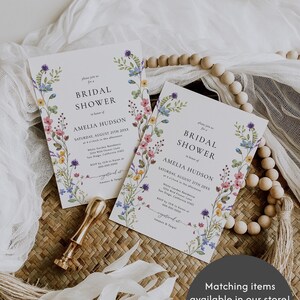 May include: Two floral bridal shower invitations with a watercolour floral border. The invitations are printed on white cardstock and feature the text "Bridal Shower" in a simple black font. The text "Amelia Hudson" is printed in a slightly larger font below the text "Bridal Shower". The invitations are laid out on a woven wicker surface with a white fabric and wooden beads.