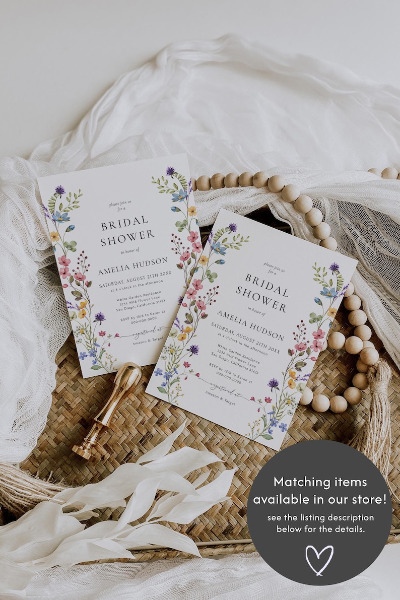 May include: Two floral bridal shower invitations with a watercolour floral border. The invitations are printed on white cardstock and feature the text "Bridal Shower" in a simple black font. The text "Amelia Hudson" is printed in a slightly larger font below the text "Bridal Shower". The invitations are laid out on a woven wicker surface with a white fabric and wooden beads.