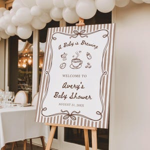 May include: A rectangular sign with brown and white vertical stripes featuring a decorative border and the text A Baby is Brewing, Welcome to Avery's Baby Shower, August 21, 20XX. The sign includes brown line art of a coffee pot, cup, and bottle.