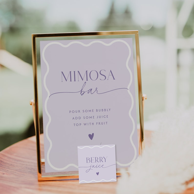 May include: A gold framed sign with a purple background and white border. The sign reads 'Mimosa bar' in a cursive font. Below the text, it says 'Pour some bubbly, add some juice, top with fruit.' A small purple card with a white border and white text that reads 'Berry juice' is in front of the sign.