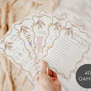 May include: A collection of baby shower game cards with a bunny theme. The cards feature illustrations of bunnies, floral accents, and various game prompts. The text "Baby Prep" and "Who Knows Mommy Best" are visible. The image includes a round graphic that says "40 GAMES".