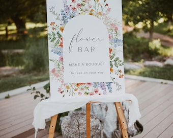 May include: A wooden easel displays a white sign with the words "Flower Bar" and "Make a Bouquet to take on your way." The sign is decorated with a floral border of colorful wildflowers. A white cloth drapes the bottom of the sign.
