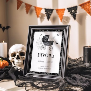 May include: A framed sign with the word "FAVORS" and a baby carriage illustration, decorated with bats and spiderwebs. The sign is part of a Halloween-themed display, with a skull, pumpkins, and bunting.