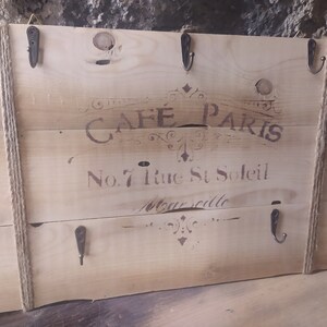 Può includere: Un cartello in legno rustico con il testo "Cafe Paris" e "No. 7 Rue St Soleil Marseille" dipinto su di esso. Il cartello ha cinque ganci in metallo ed è decorato con spago.