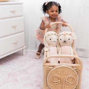 May include: A toddler in a floral dress pushes a light-colored wooden wagon containing two plush bunny toys. The wagon has a circular cutout on the front and is made of woven material. The bunnies are light pink with floral headbands.