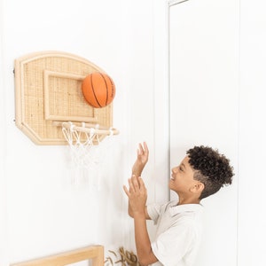 May include: A child plays with a miniature basketball hoop. The hoop is made of light-colored wood and woven material. An orange basketball is in mid-air, heading towards the net. The child is wearing a light-colored polo shirt and khaki shorts.