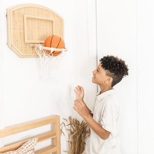 May include: A wall-mounted basketball hoop with a woven backboard and a white net. An orange basketball is in the net. The hoop is mounted on a white wall. A young person is standing nearby.