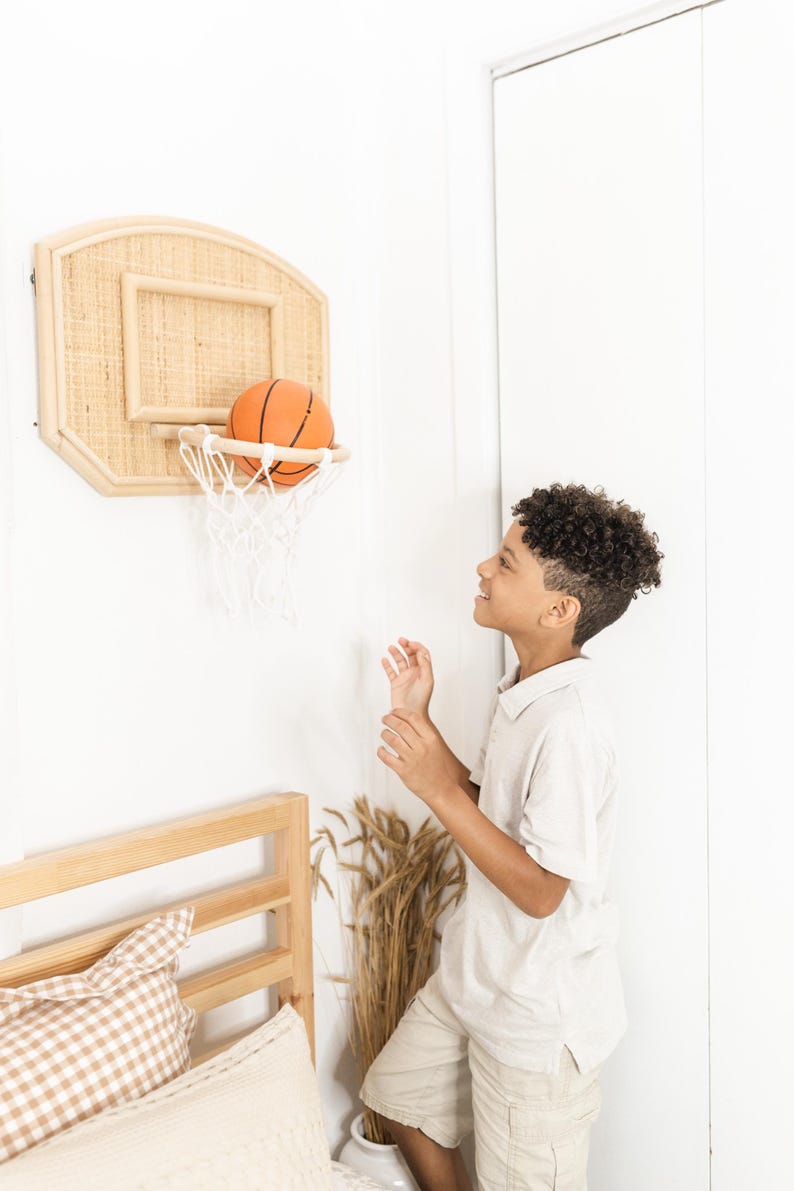 May include: A wall-mounted basketball hoop with a woven backboard and a white net. An orange basketball is in the net. The hoop is mounted on a white wall. A young person is standing nearby.