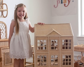 May include: A large, three-story dollhouse made of woven natural material. The dollhouse has multiple windows and a pitched roof. A young girl in a white dress stands next to the dollhouse. A banner in the background reads "Oh Yay Let's Play!"