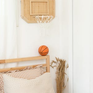 May include: A woven rattan basketball hoop with a white net is mounted on a white wall. An orange basketball sits on a wooden bed frame, next to a beige pillow and a vase of dried wheat. The room has a neutral color palette.
