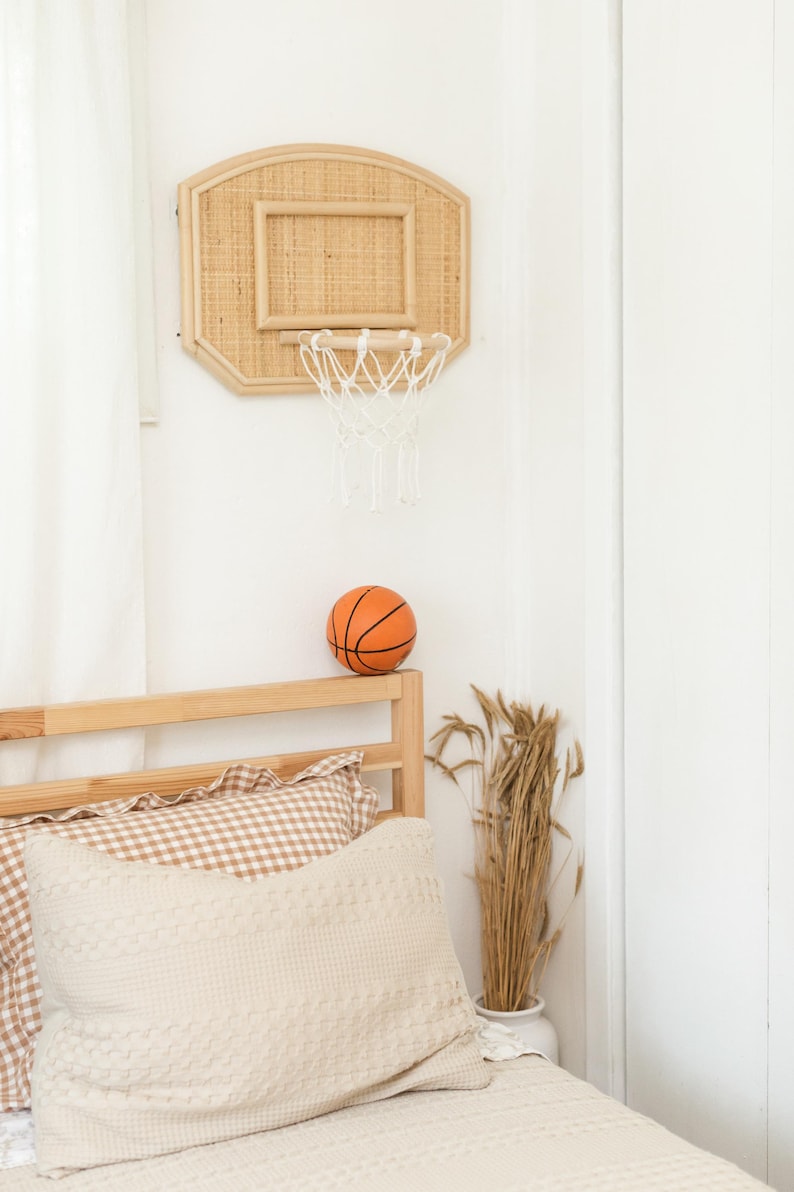 May include: A woven rattan basketball hoop with a white net is mounted on a white wall. An orange basketball sits on a wooden bed frame, next to a beige pillow and a vase of dried wheat. The room has a neutral color palette.
