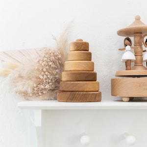 May include: A wooden stacking toy with six rings in various sizes, arranged in a pyramid shape on a white shelf. The toy is made of natural wood and has a rustic finish. A wooden music box with a carousel design is visible in the background.