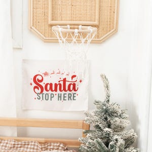 May include: A decorative basketball hoop made of woven material hangs on a white wall. Below it, a banner reads "Santa Stop Here" in red and green. A small, snowy Christmas tree sits in a white vase.