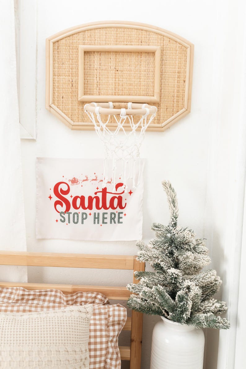 May include: A decorative basketball hoop made of woven material hangs on a white wall. Below it, a banner reads "Santa Stop Here" in red and green. A small, snowy Christmas tree sits in a white vase.