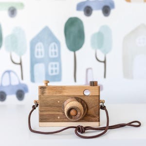 May include: A wooden toy camera with a brown strap. The camera has a round lens and is made of light and dark wood. The camera is in focus and the background is blurred.