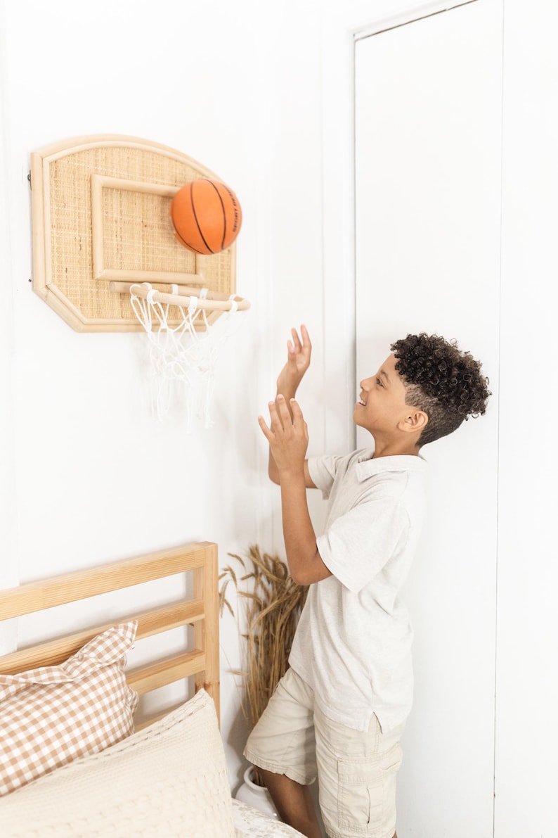 May include: A child plays with a miniature basketball hoop. The hoop is made of light-colored wood and woven material. An orange basketball is in mid-air, heading towards the net. The child is wearing a light-colored polo shirt and khaki shorts.
