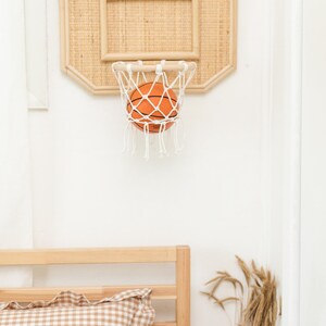May include: A miniature basketball hoop with a woven backboard and a white net, holding an orange basketball. The hoop is mounted on a white wall. The overall theme is sports and home decor.