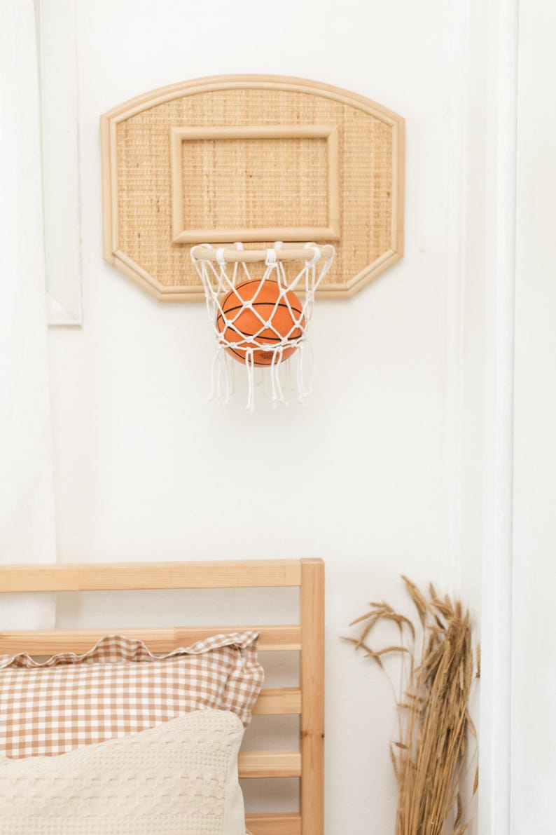 May include: A miniature basketball hoop with a woven backboard and a white net, holding an orange basketball. The hoop is mounted on a white wall. The overall theme is sports and home decor.