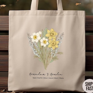 May include: Beige tote bag featuring a floral design with yellow and white daffodils and lavender. The text "Grandma's Garden" is below the flowers, with names listed underneath. The bag has cream-colored handles.