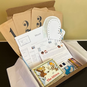 Op de afbeelding: Een bruine kartonnen doos gevuld met een kinderboek getiteld "The Mitten" van Jan Brett, een witten wanten knutselwerk, nummerkaarten, kleurpotloden en andere educatieve materialen. De doos is gelabeld "Read Play Grow."