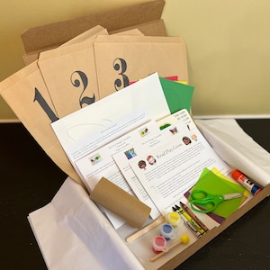 May include: A cardboard box filled with craft supplies for a children's activity. The box contains paper, paint, crayons, scissors, glue, and a cardboard tube. The box also includes instructions for the activity, which is titled "Read, Play, Grow".