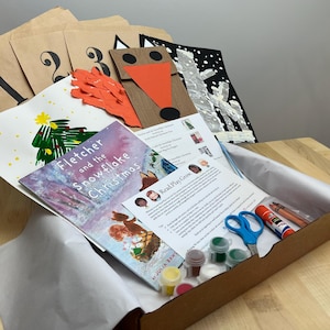 May include: A craft kit with a book titled "Fletcher and the Snowflake Christmas" and art supplies. The kit includes paint, scissors, glue, crayons, and numbered paper bags. A drawing of a Christmas tree is visible.