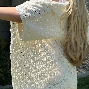 May include: A white crochet cardigan with a delicate openwork pattern. The cardigan has a relaxed fit and three-quarter sleeves.