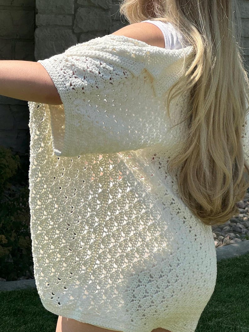 May include: A white crochet cardigan with a delicate openwork pattern. The cardigan has a relaxed fit and three-quarter sleeves.