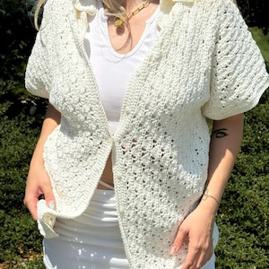 May include: A white crochet button-up shirt with short sleeves. The shirt has a delicate open-weave pattern.