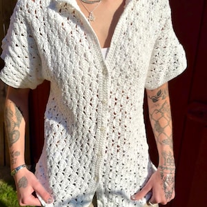 May include: White crochet button-up shirt with short sleeves. The shirt has a delicate openwork pattern.