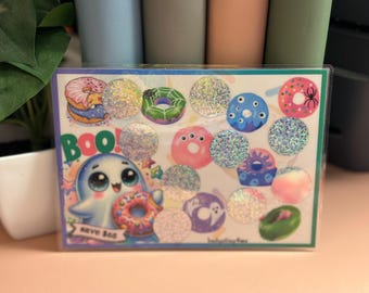May include: A colorful card with a ghost holding a donut and the word "BOO!" printed on it. The card is decorated with glitter and has a variety of donut-shaped stickers with different designs, including eyes, spiders, and ghosts.