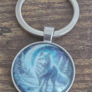 May include: A circular silver-toned keychain featuring a domed glass cabochon insert. The insert displays a graphic illustration of a wolf standing in a snowy forest landscape under a large, bright crescent moon in shades of blue and white.