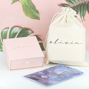 May include: A pink gift box with the word "olivia" printed on the top, a white drawstring bag with the word "olivia" printed on the front, and a blue and white card with the word "diamond" printed on it.
