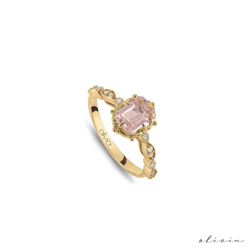 May include: A gold ring with a pink gemstone and small diamonds set in a delicate, ornate design. The ring is inscribed with the word "olivia".