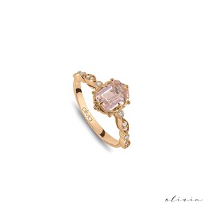 May include: A delicate rose gold ring with a pink gemstone surrounded by smaller diamonds. The band is adorned with a twisted design and features the word "olivia" engraved on the side.