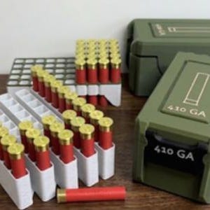 May include: A collection of red and gold 410 GA shotgun shells arranged in white plastic organisers, accompanied by two olive green storage cases. Each case features a moulded graphic of a shell and the text 410 GA printed on the front lid.