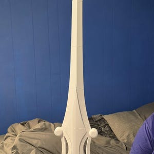 May include: A white sword with a long, pointed blade and a decorative hilt. The hilt features rounded accents. The sword is held against a blue wall and a gray blanket.