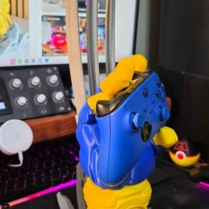 May include: A blue and yellow controller stand shaped like a gloved hand with three long, silver-colored claws extending upward. The stand features a sculpted base and holds a blue wireless game controller securely within its grasp.